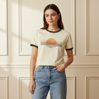 Woman wearing a t-shirt with a sunset design and text, standing against a beige paneled wall.