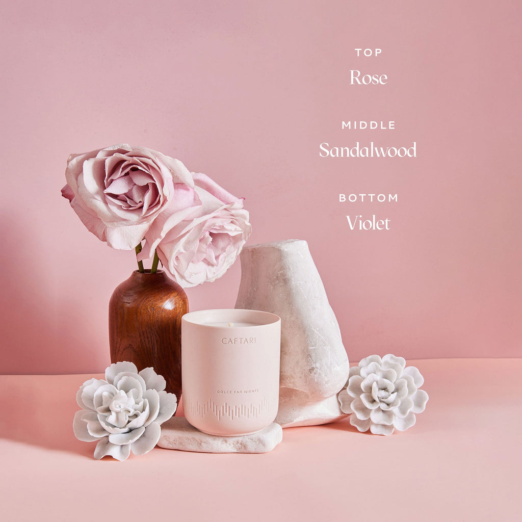 Caftari Dolce Far Niente rose candle with sandalwood and violet fragrance notes in a soft pink floral setting