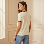 Woman wearing a beige t-shirt with green trim and blue jeans in a room with beige walls.