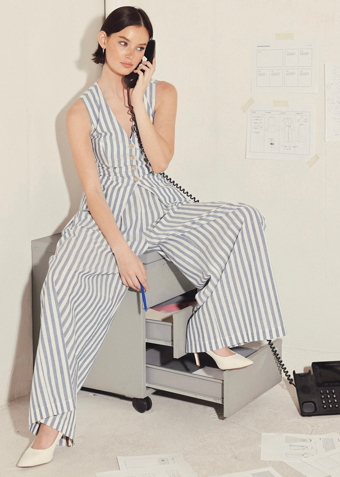 Woman in a striped jumpsuit sitting on a rolling chair, talking on a phone in an office setting. Main image
