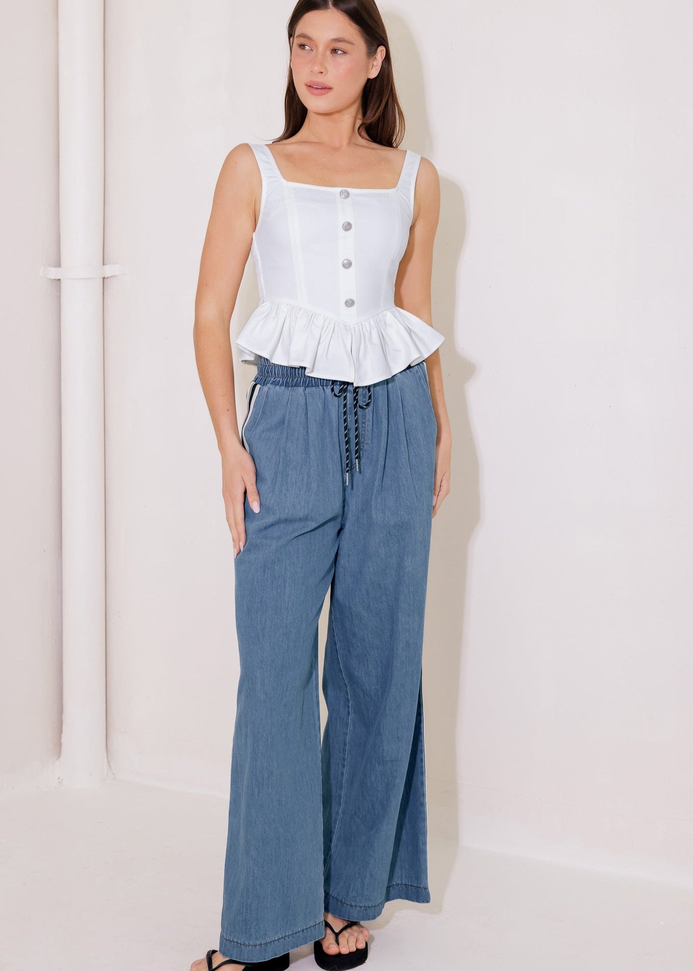 Woman wearing a white sleeveless top with ruffled details and blue wide-leg pants on a plain background